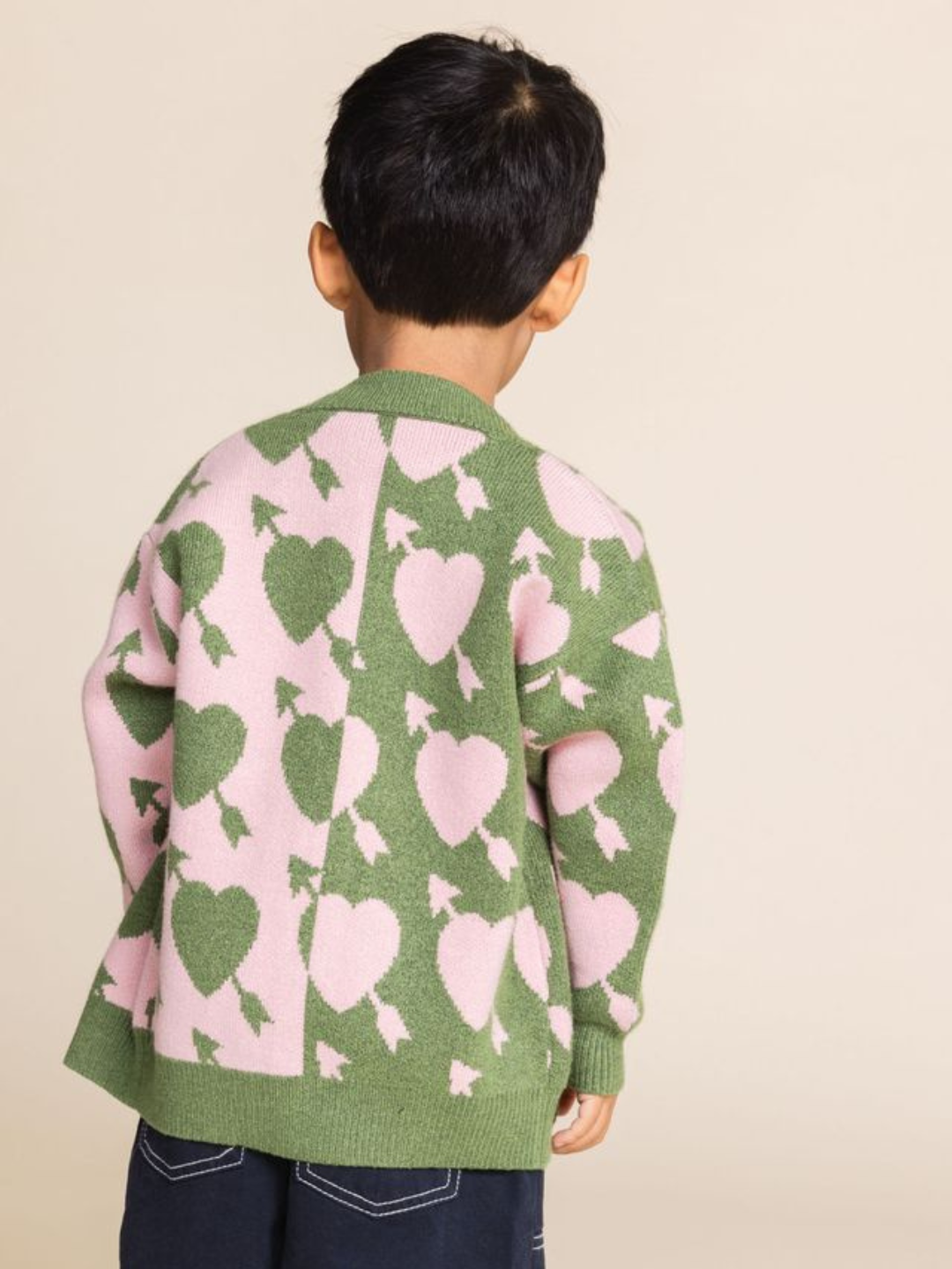 A child stands facing away, sporting the LOVESTRUCK CARDIGAN in green and pink, adorned with a heart and arrow pattern, perfect for cozy photos.