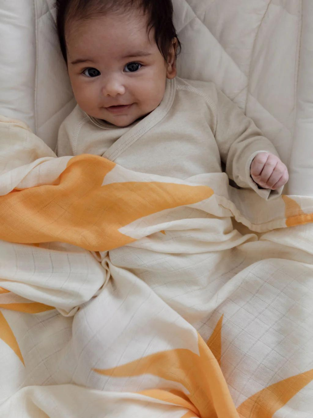 A baby wearing a light-colored outfit is lying down, partially covered by an ORGANIC BAMBOO SWADDLE WRAP with a cream and orange floral pattern.