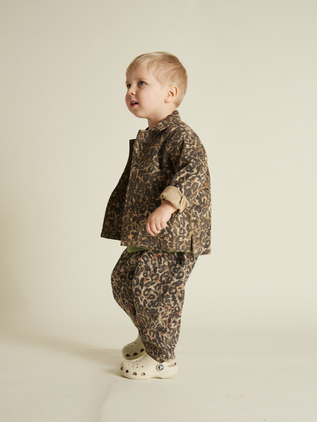 A child stands sideways, modeling an oversized leopard-print FEN LEOPARD JACKET made of organic cotton, paired with white clogs, against a plain background.