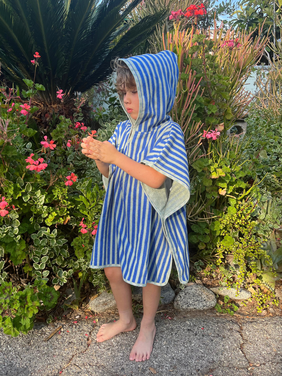 Green | Child wearing a blue and white striped hooded towel standing in front of greenery with pink flowers.