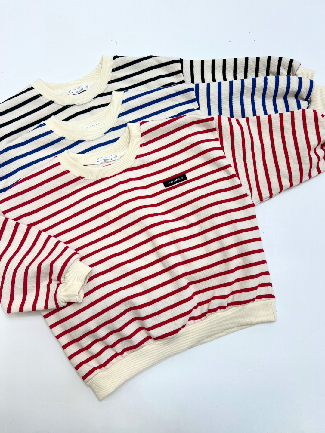 Three BRETON BABY SWEATSHIRTs are stacked on a white surface: the top has bold red stripes, the middle classic blue, and the bottom sleek black stripes.
