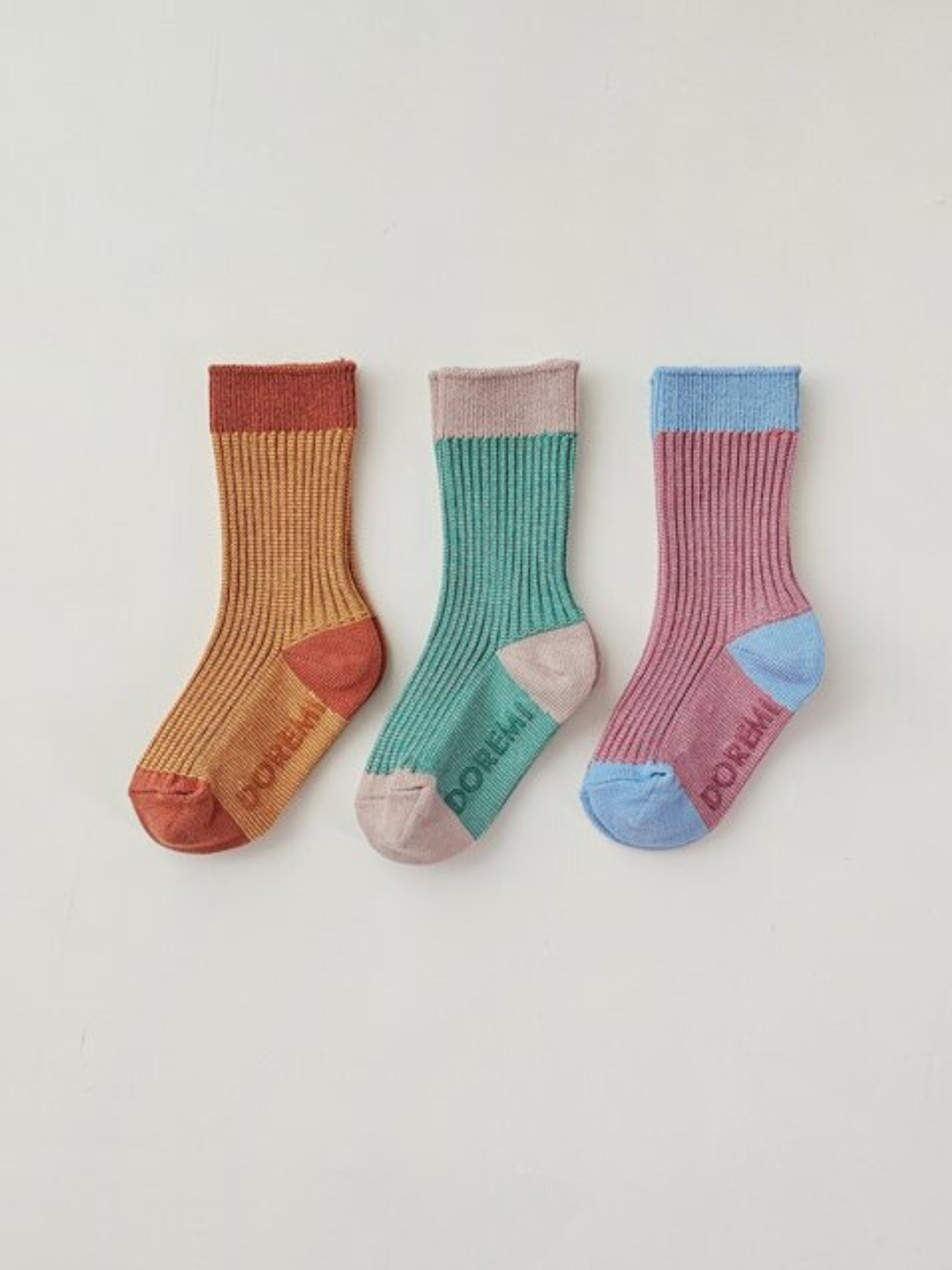 Three pairs of colorful ribbed socks from the TRIO SOCK SET laid out side by side on a white surface, showcasing vibrant color combinations: red and orange, green and pink, and purple and blue.