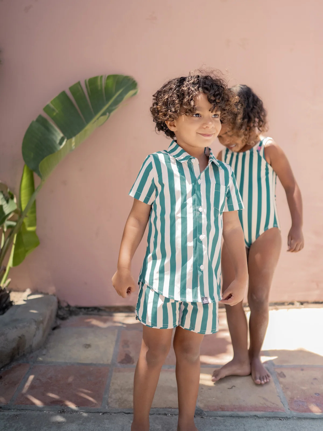 Charleston | Two children wear RETRO FLARE BOARDSHORTS CABANA STRIPE—green and white striped swim shorts made from recycled polyester—as they stand on a tiled patio in front of a pink wall with a large green plant.