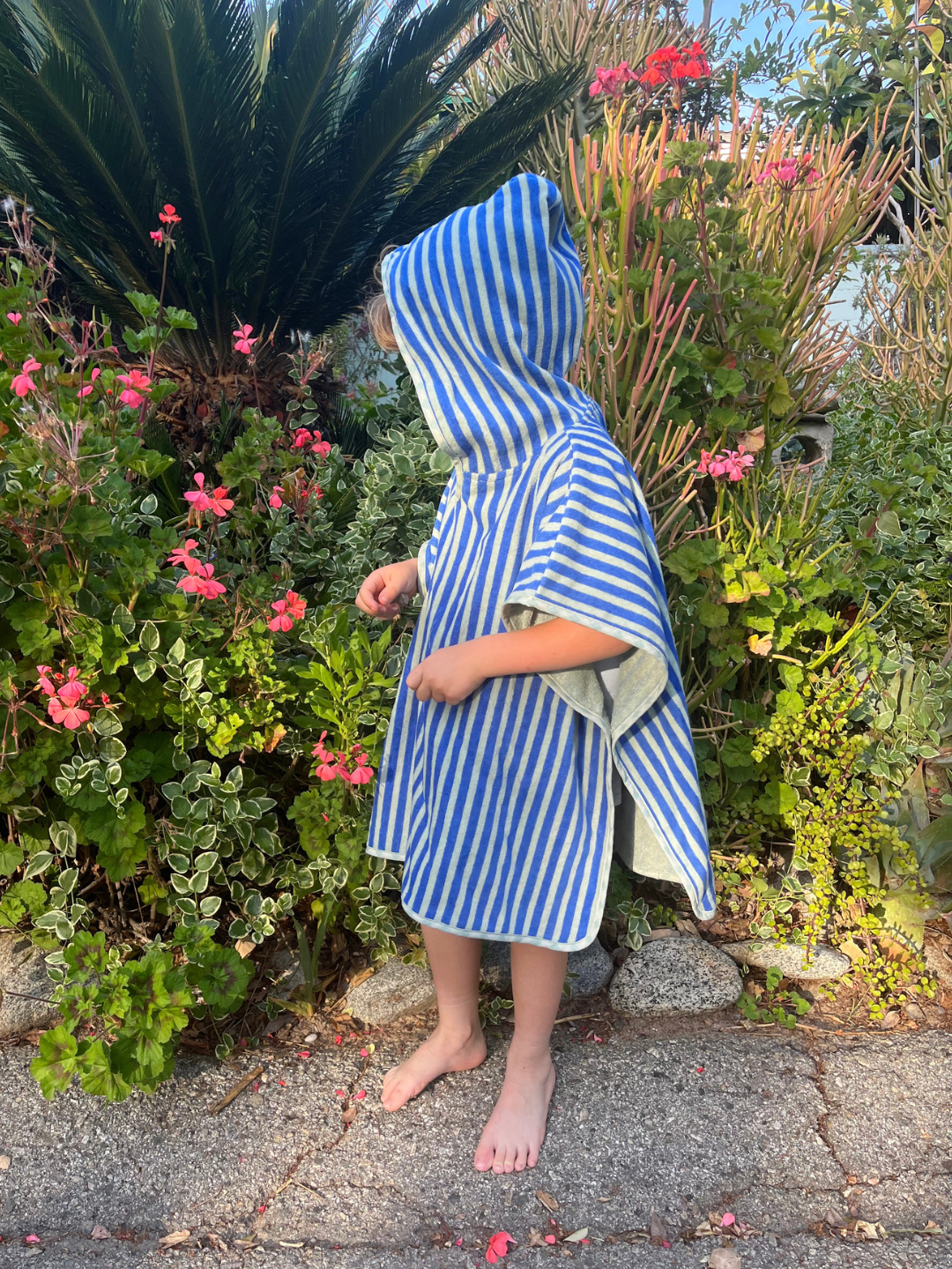 Child wearing a blue and white striped hooded towel standing in front of greenery with pink flowers.
