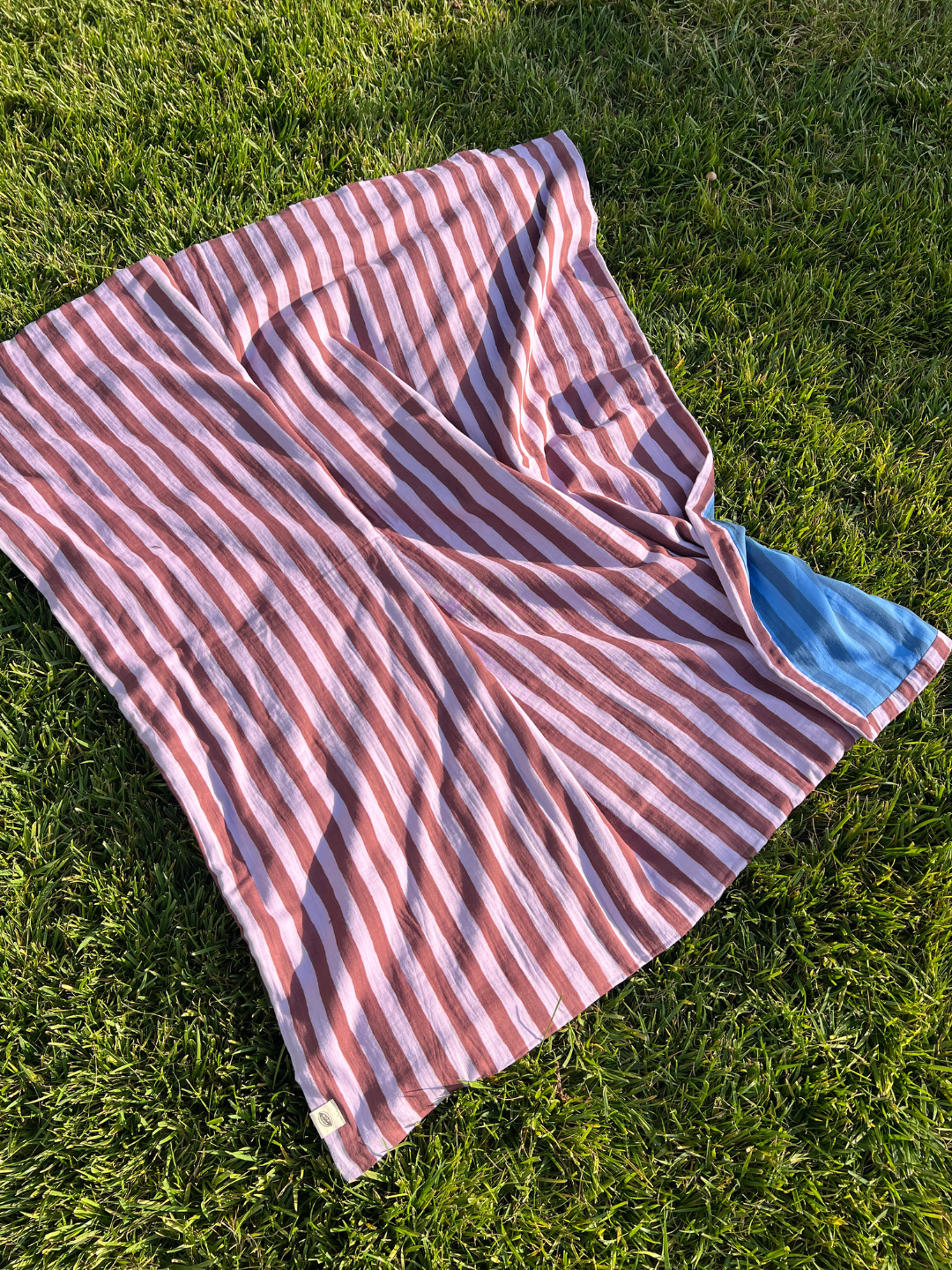 Purple Stripe | The organic cotton quilt in purple and maroon stripes, is shown on grass, with its blue striped underside showing.