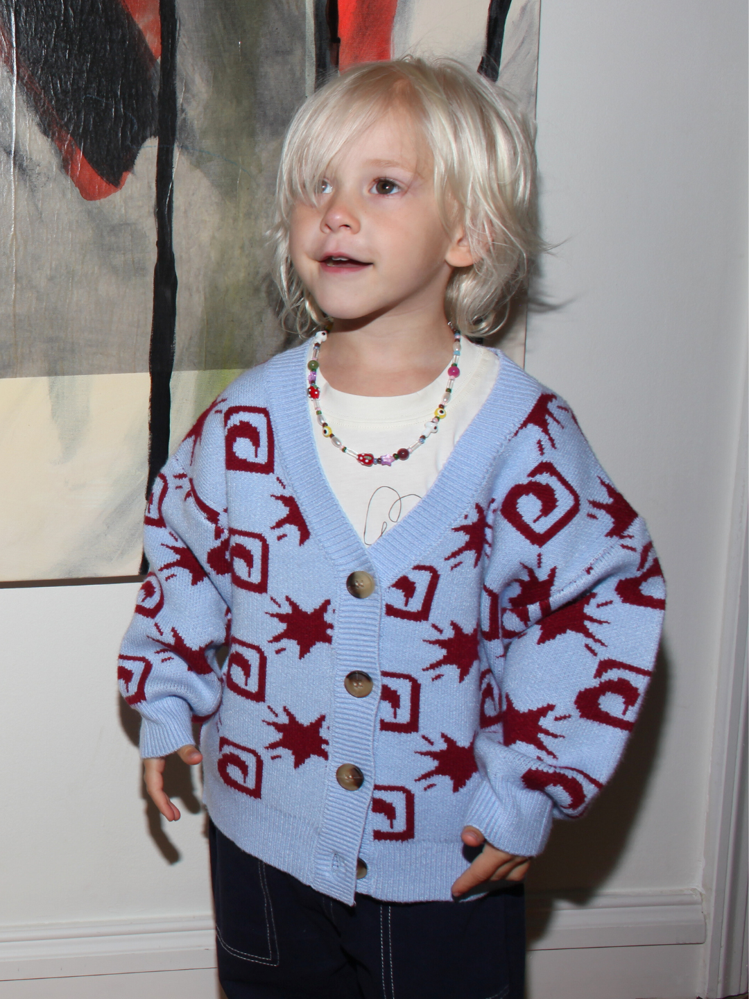 A young child with blond hair wears a blue and red patterned cardigan, a white shirt, and the GAIA NECKLACE—beaded with handmade strawberry and mushroom beads—while standing indoors near abstract wall art.