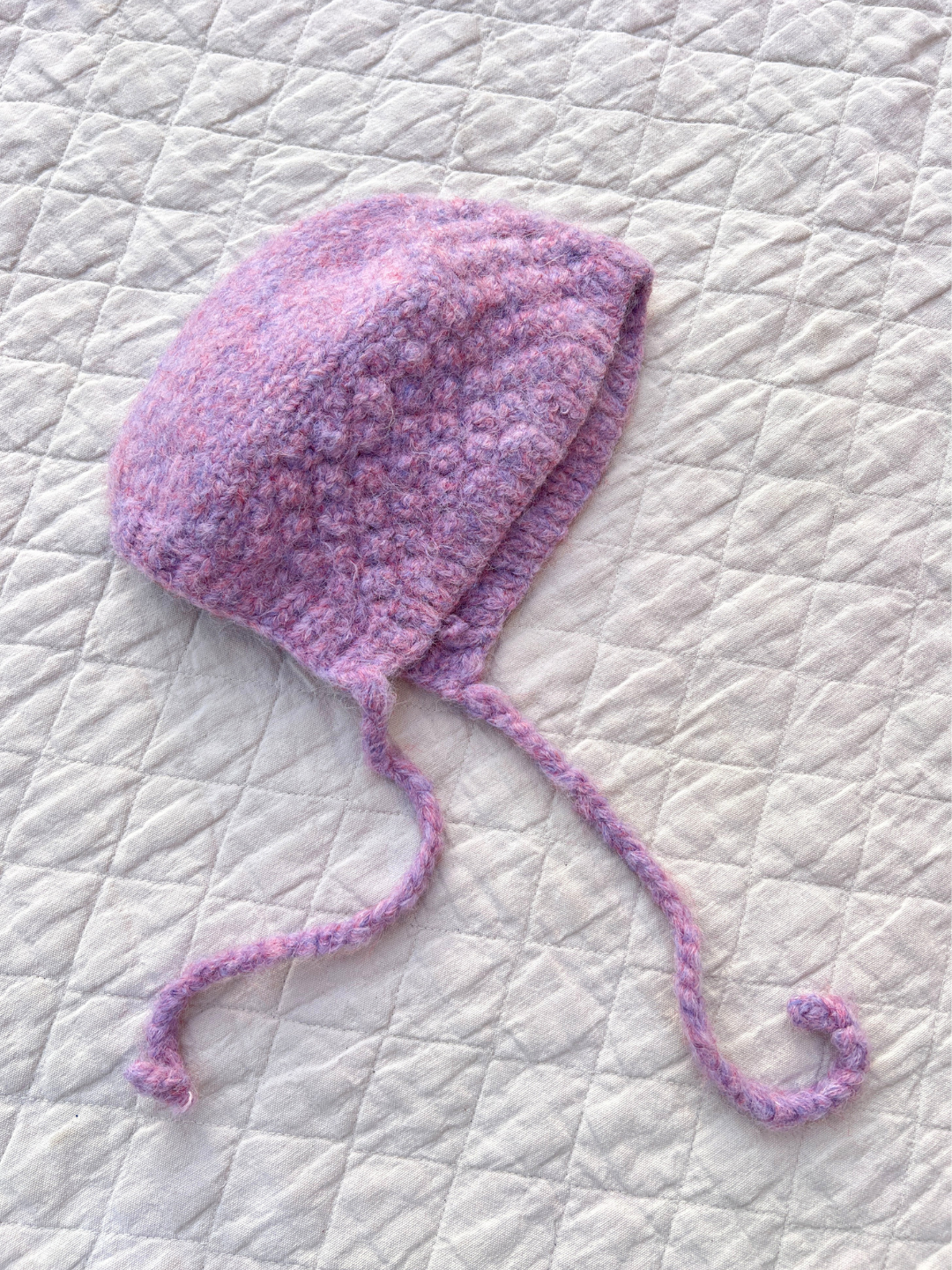 The POPCORN BONNET in a cozy purple knit features two curly ties and rests elegantly on a quilted white background, exuding its felted texture charm.