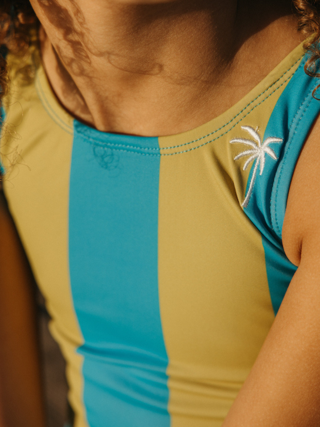 Kelp | A child is wearing a Retro Stripe Swimsuit in Kelp. The close up in on the Palm Tree embroidery. 