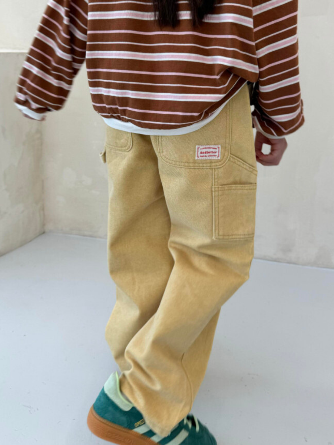 Ochre | A child wearing a striped long-sleeve shirt, PIGMENT CARPENTER JEANS with an elastic waist, and green shoes is walking indoors. The image is taken from behind.