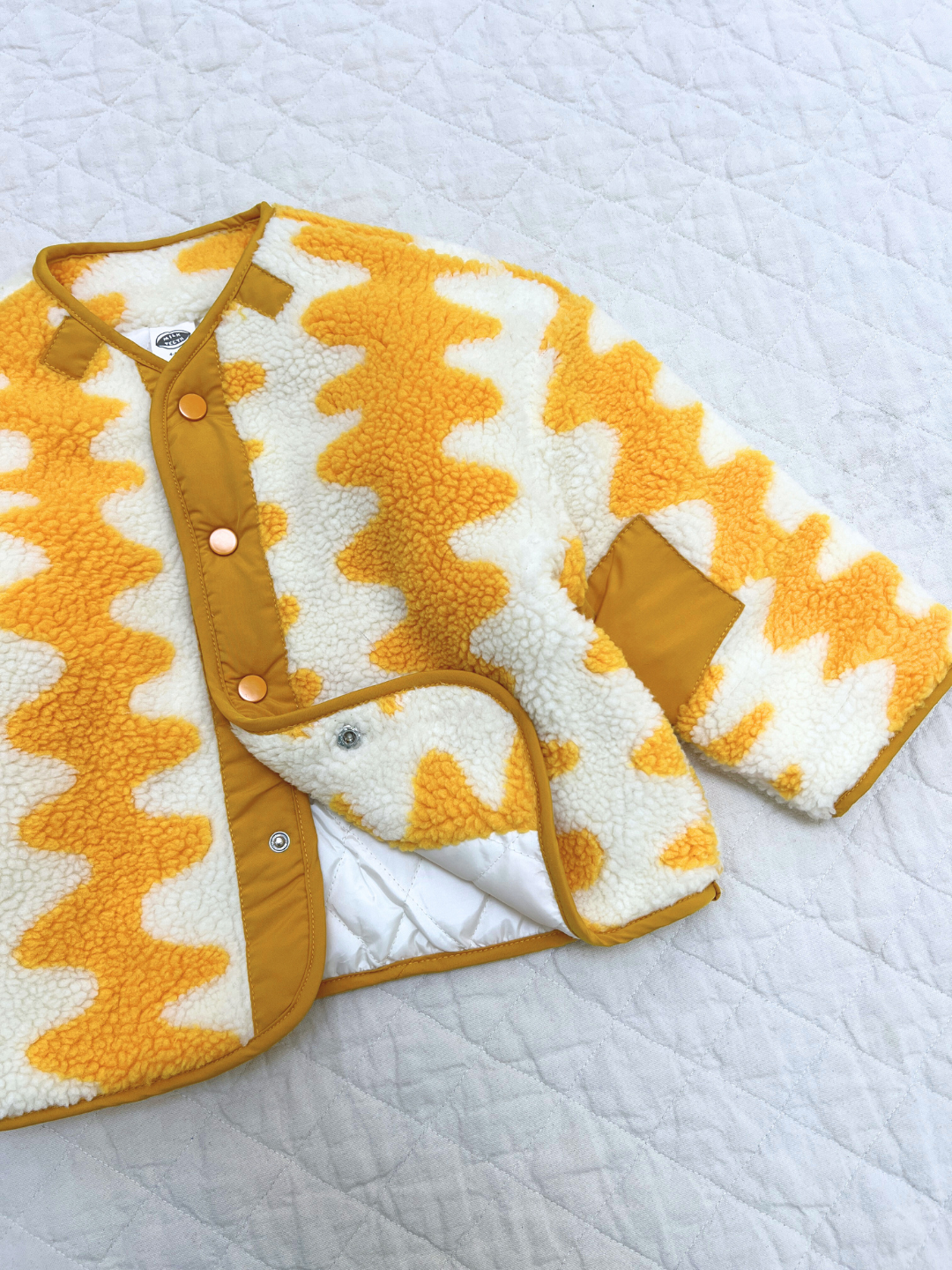 The WAVELENGTH SHERPA JACKET, featuring a yellow and white zigzag pattern and snap buttons, is displayed on a white quilted surface.