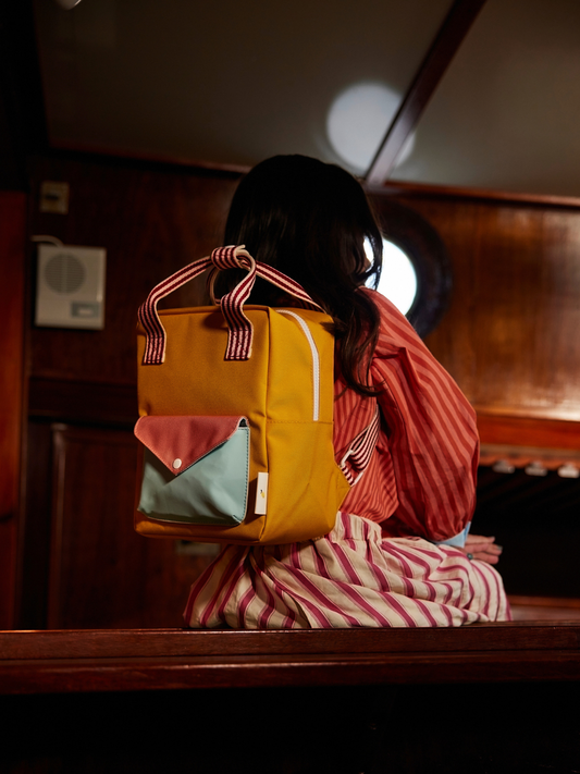 Second image of Submarine Yellow | The SMALL ENVELOPE BACKPACK is a compact yellow bag made from recycled PET bottles, with red and white striped handles and a light blue front pocket with a pink flap, shown on a plain white background.