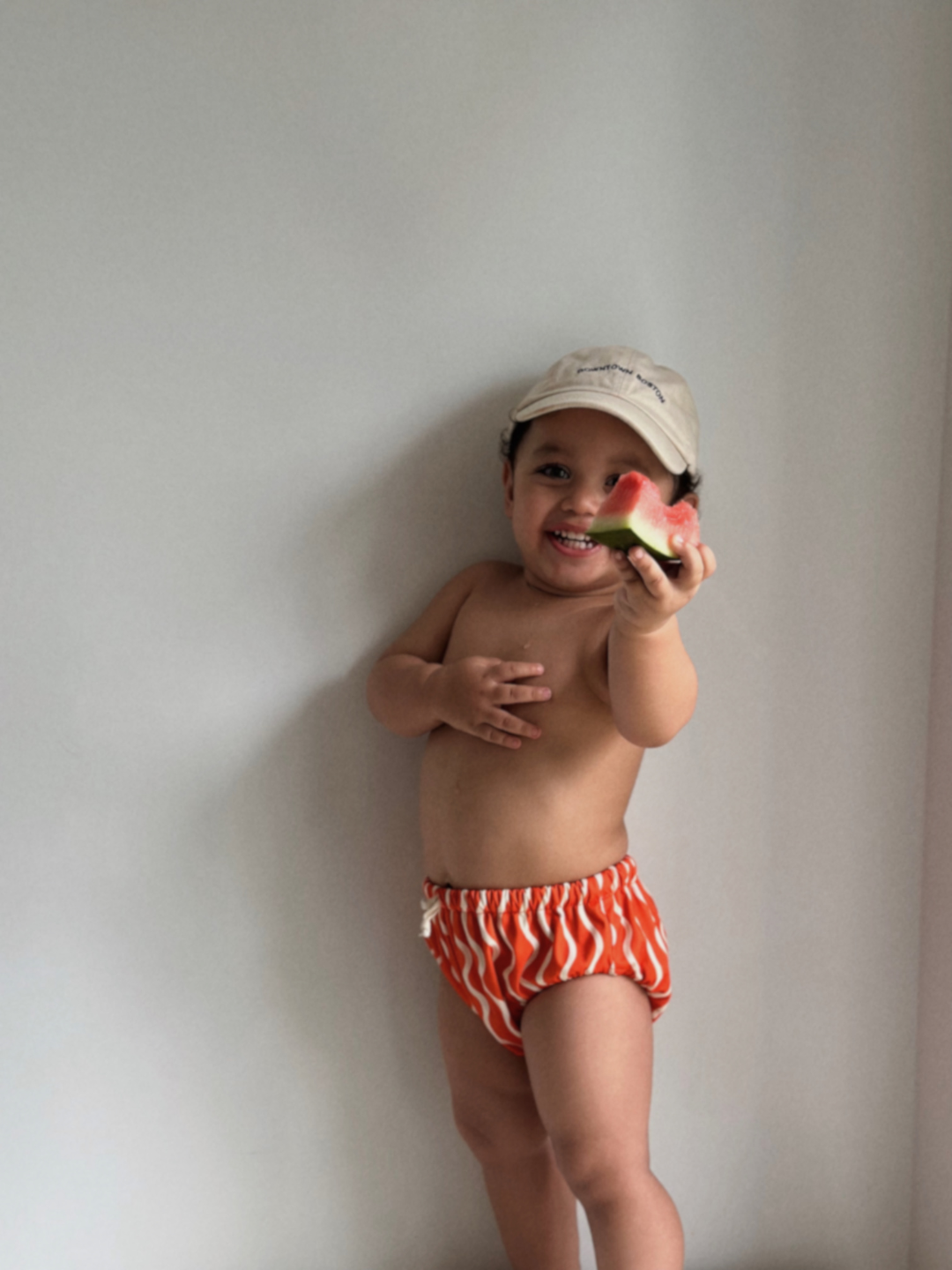 The front view of the swim diaper with an elastic waist with a tie and elastic leg holes on a child standing up. The diaper is a bright orange with wavy and vertical cream colored lines.