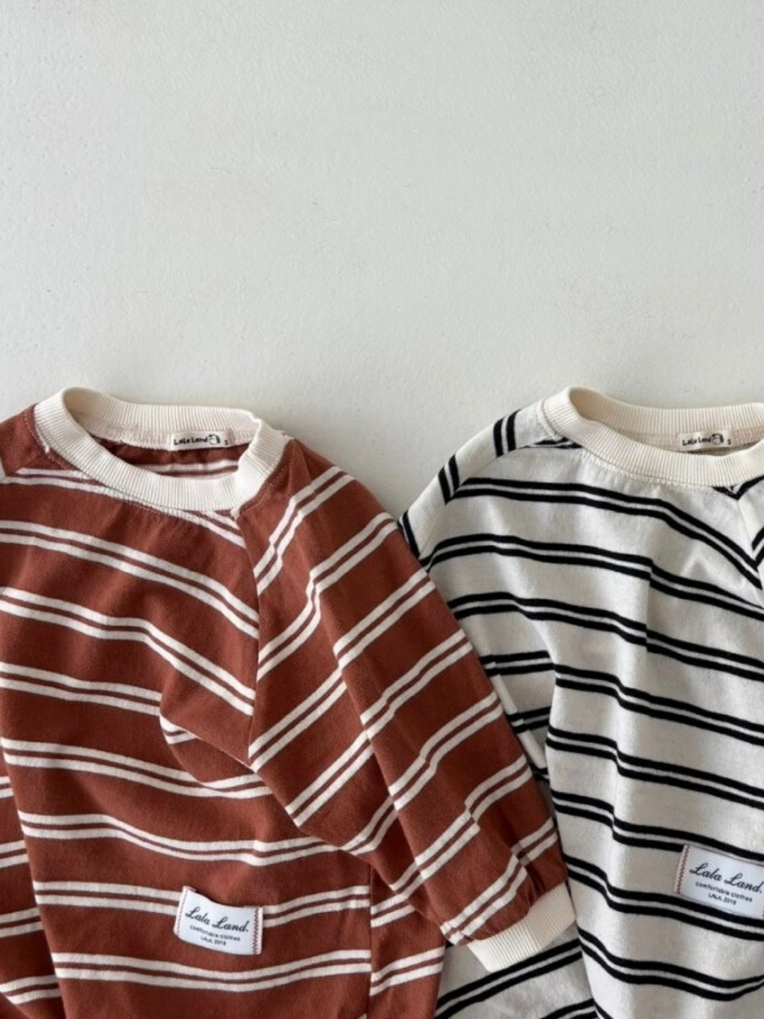 The SLOUCHY STRIPE ROMPER is a cozy staple from a popular Korean baby brand—two long-sleeve rompers in soft cotton jersey, one brown with white stripes and one white with black stripes, both featuring a label on the bottom hem.