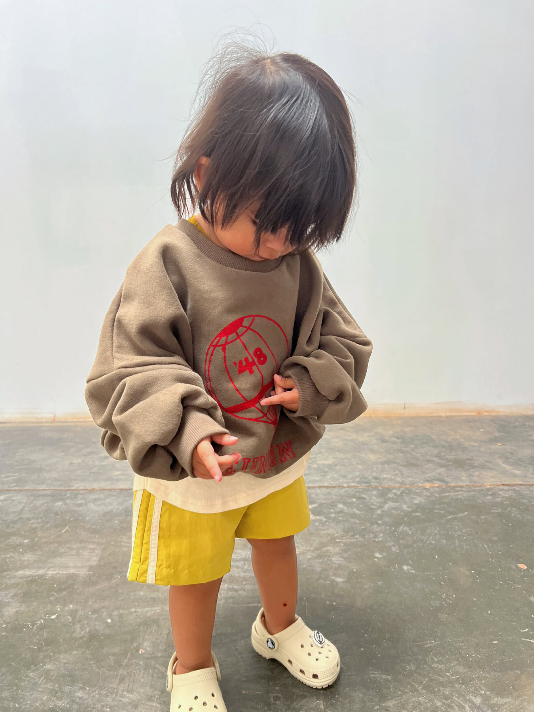 Olive | A young child stands indoors, curiously looking down at their REUNION SWEATSHIRT—an oversized brown top with a playful red graphic—paired with yellow shorts and light slip-on shoes.