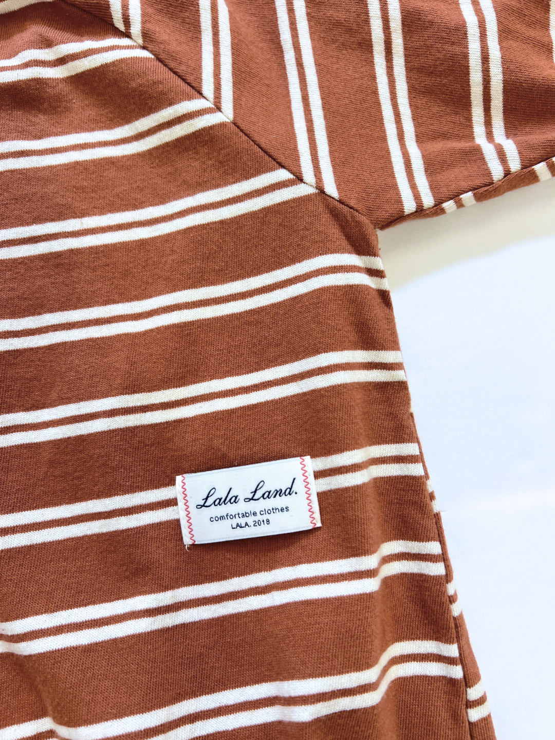 The SLOUCHY STRIPE ROMPER, a brown and white striped piece from a Korean baby brand, is made from soft cotton jersey and features a sewn label reading "Lala Land. comfortable clothes LALA 2019" in black and red text.