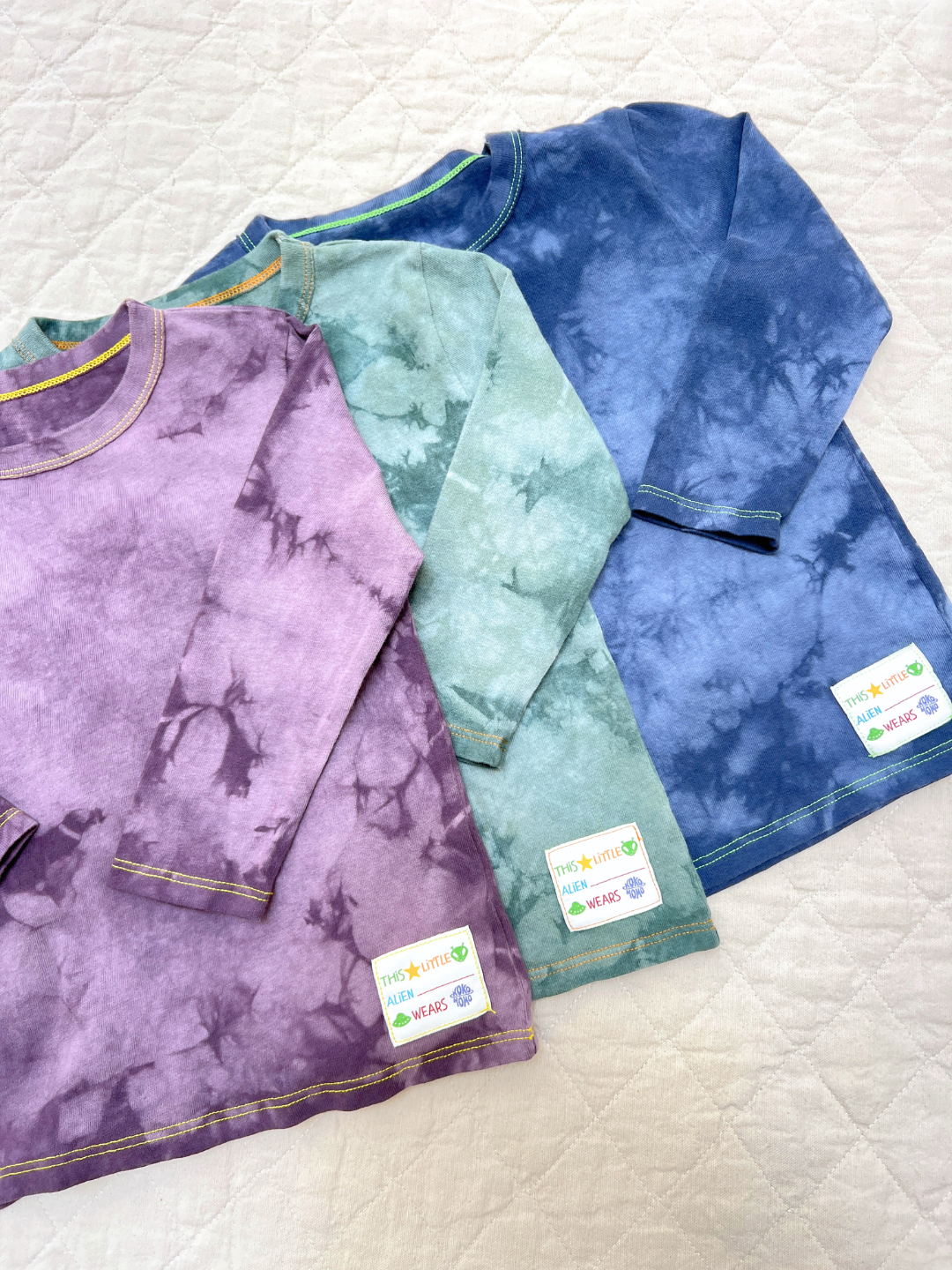 Three COSMO TIE DYE LONG SLEEVE shirts in purple, green, and blue are laid out on a quilted surface, each with a white label near the hem—perfect for adding LA flair to your wardrobe.