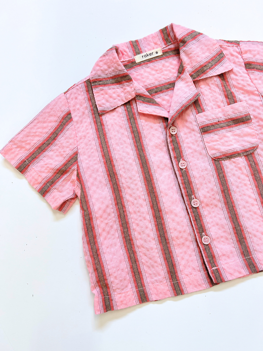 Second image of The HAVANA STRIPE SHIRT is a vintage-inspired, short-sleeved pink cotton shirt with vertical red, black, and white stripes and a single chest pocket, shown laid flat against a white background.