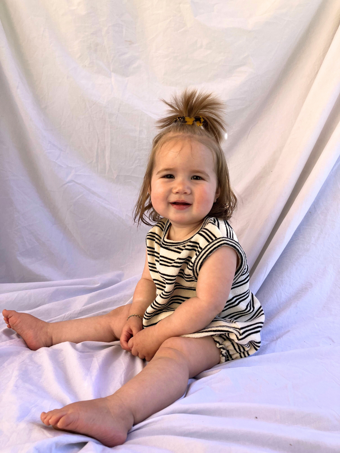 Stripe | A toddler wearing the Terry Bubble Bodysuit in cream terrycloth printed with black stripes. She wears a high ponytail and a gold bracelet, and is seated on a beige cloth backdrop.