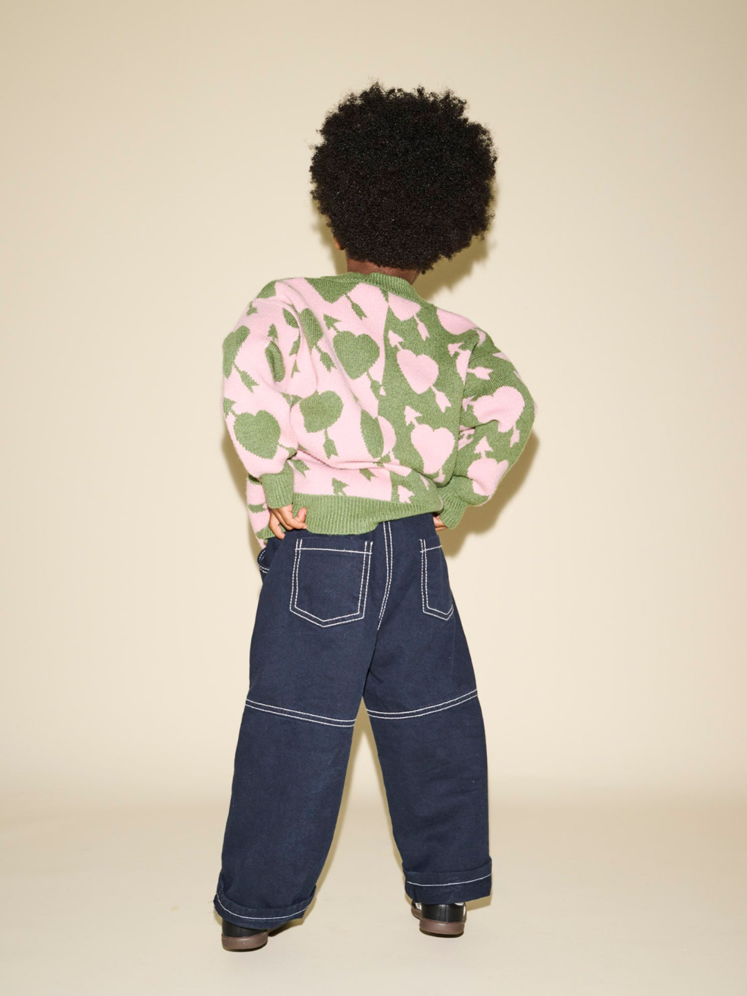 Person standing with their back to the camera, wearing the LOVESTRUCK CARDIGAN featuring a green and pink heart pattern, paired with blue wide-leg jeans and black shoes. The description captures every detail perfectly for photos or measurements.