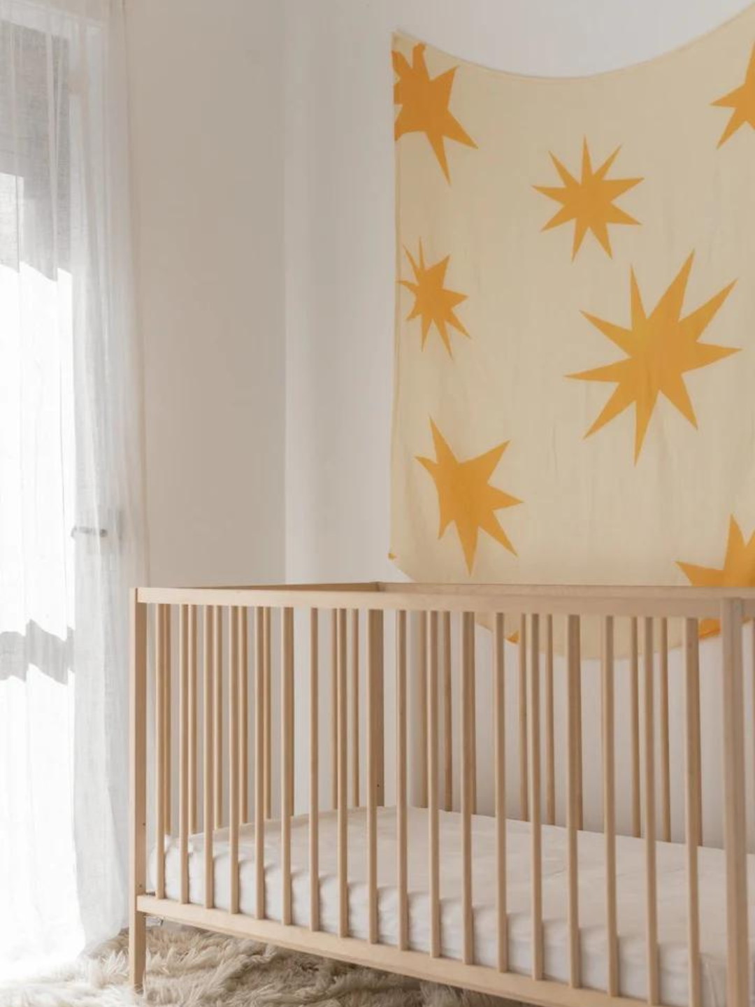 A light-colored, minimalist nursery features a wooden crib with a white mattress and a wall hanging adorned with large yellow stars. Sheer curtains gently filter the natural light into the room. Covering the floor is a beige fluffy rug, while ORGANIC BAMBOO SWADDLE WRAPs lay ready for use near the crib.