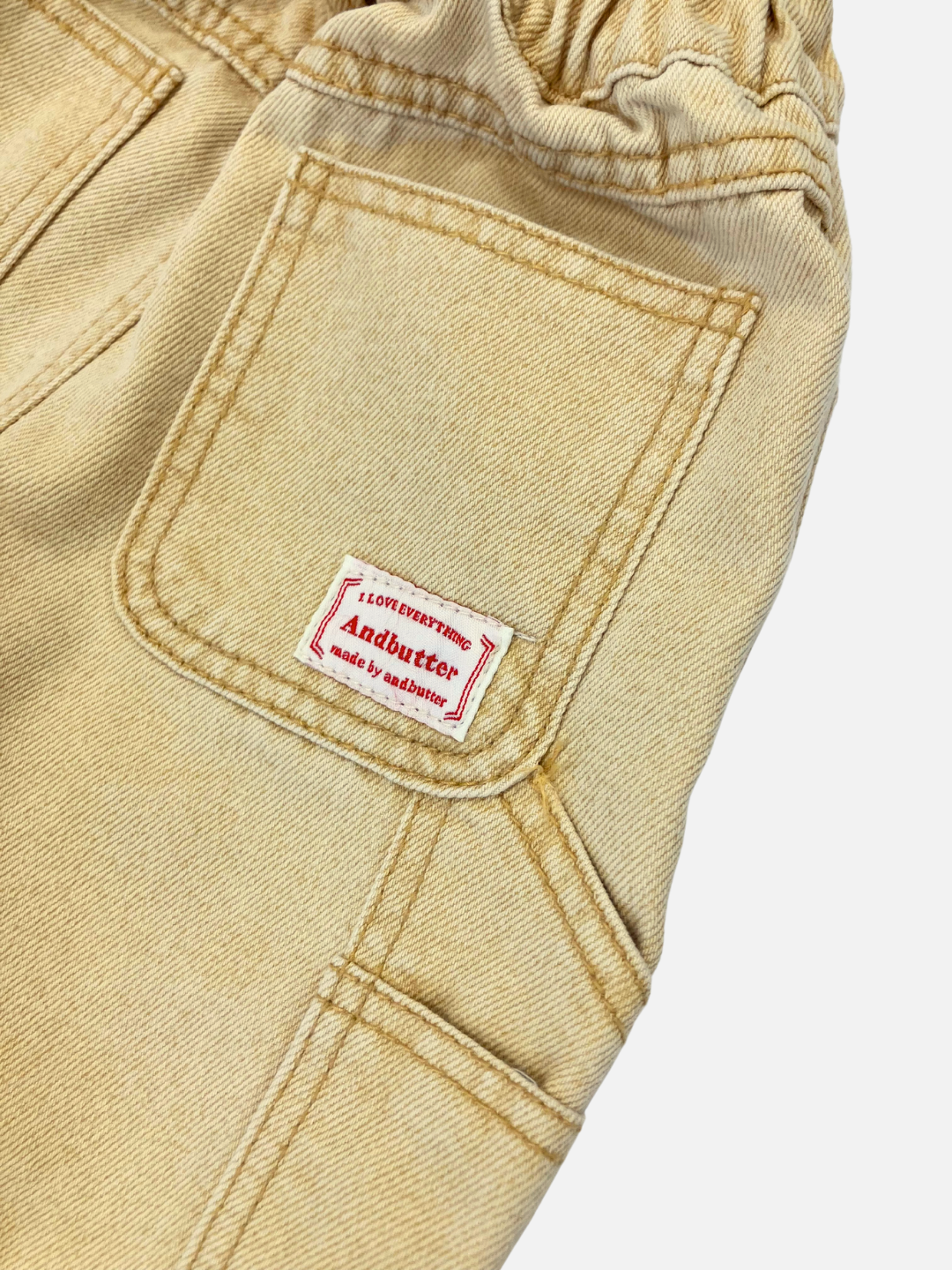 Ochre | Close-up of the beige Pigment Carpenter Jeans fabric with contrasting stitching, showcasing a small red and white label that reads "And butter" above the pocket.