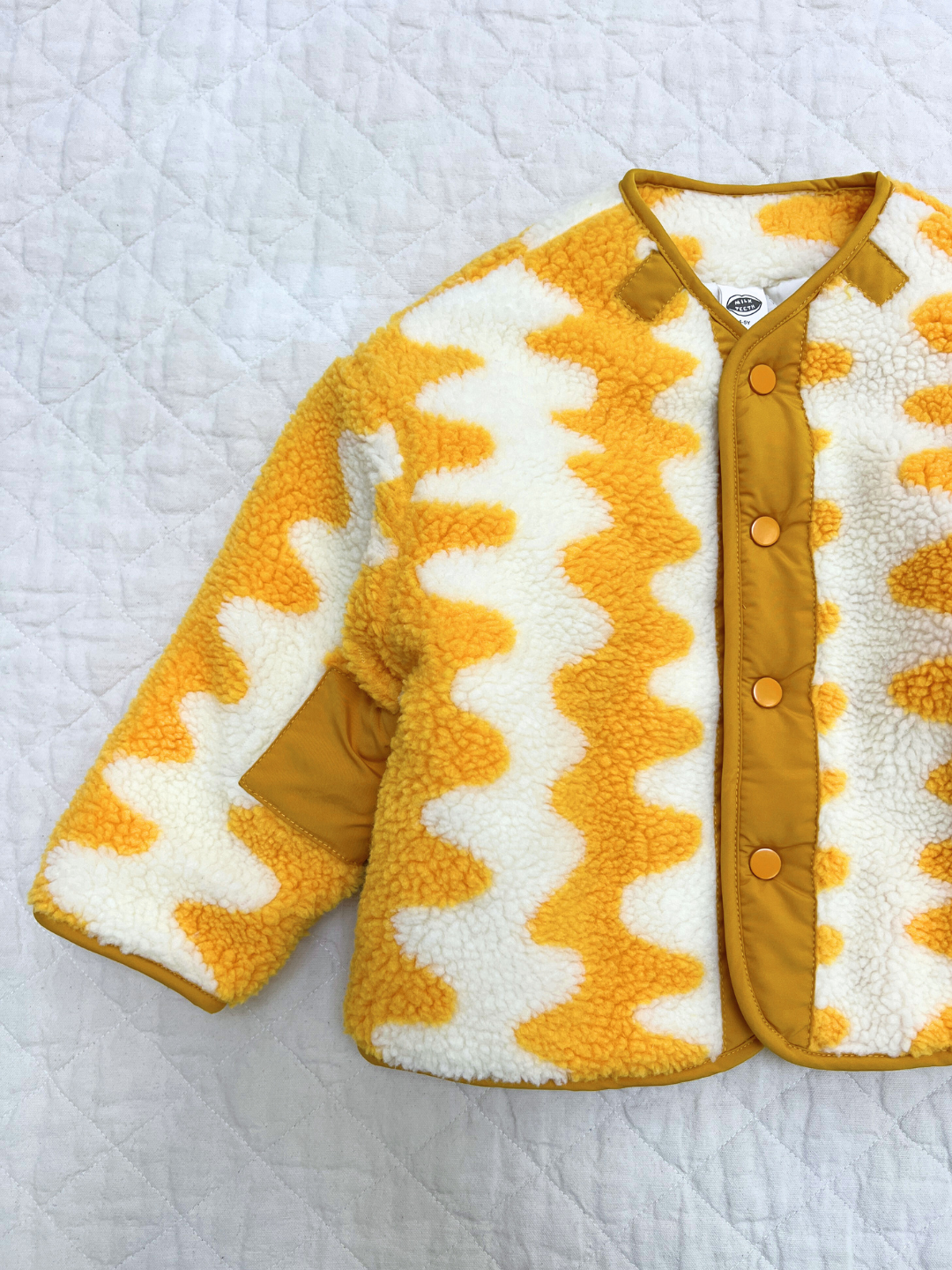 The WAVELENGTH SHERPA JACKET is a children's fuzzy fleece with a yellow and white zigzag design, yellow trim, and yellow snap buttons, displayed flat on a quilted white surface.