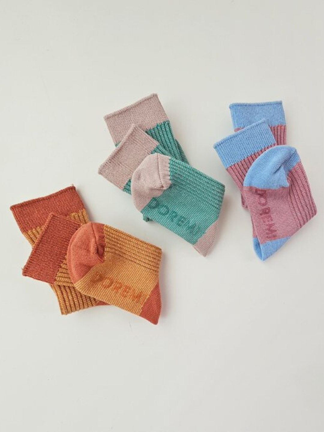 Three pairs from the TRIO SOCK SET are arranged on a white surface. The colorful ribbed socks come in shades of red and orange, green and pink, and blue and purple, showcasing the vibrant palette characteristic of this popular Korean brand.