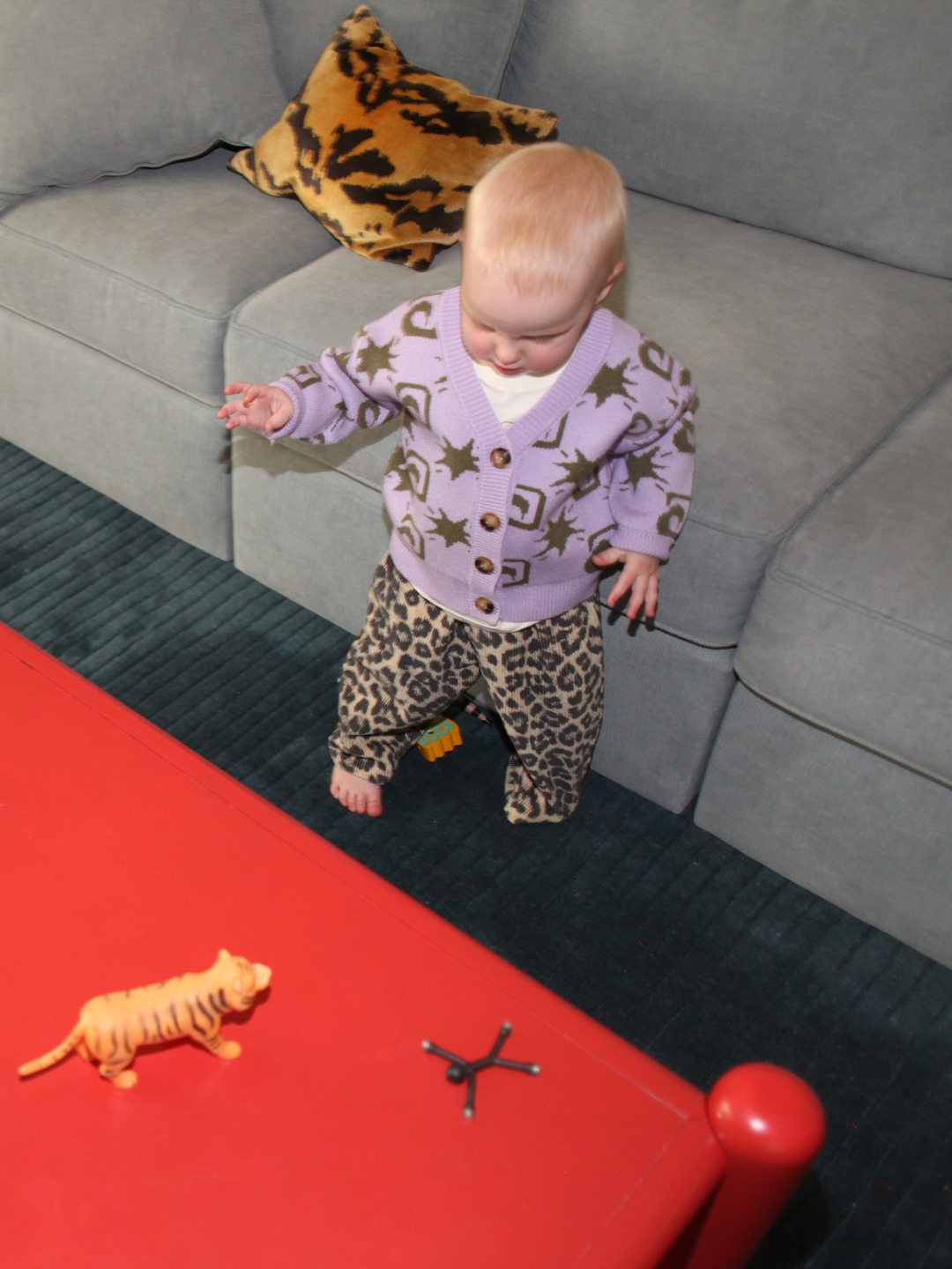 Baby in a purple cardigan and leopard print pants standing on a red surface with toys.