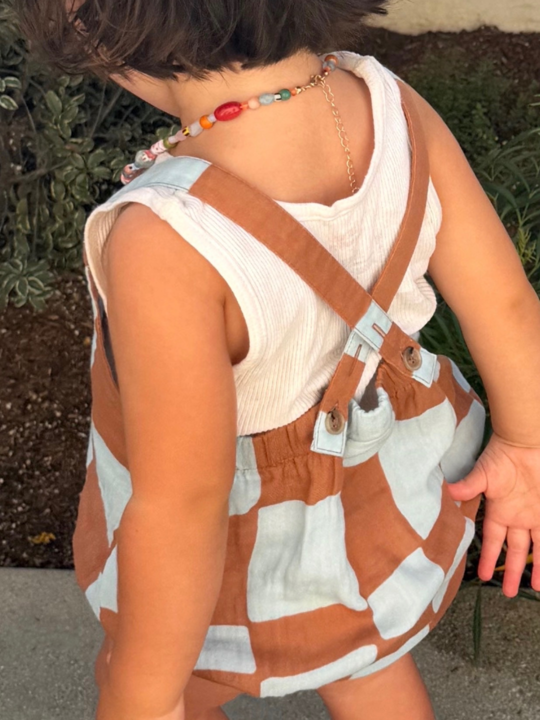 Blue Checkers | back view of child wearing a blue and brown checkerboard romper with crossback straps, over a white top outdoors.