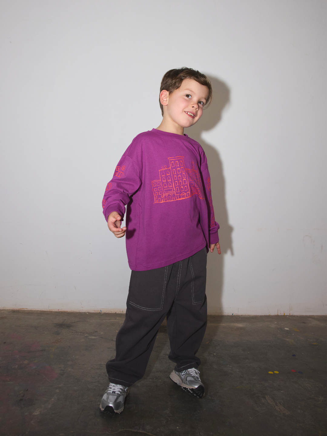 Charcoal | Child wearing baggy dark grey pants with a magenta longsleeve tee, standing on a concrete floor in front f a white wall.