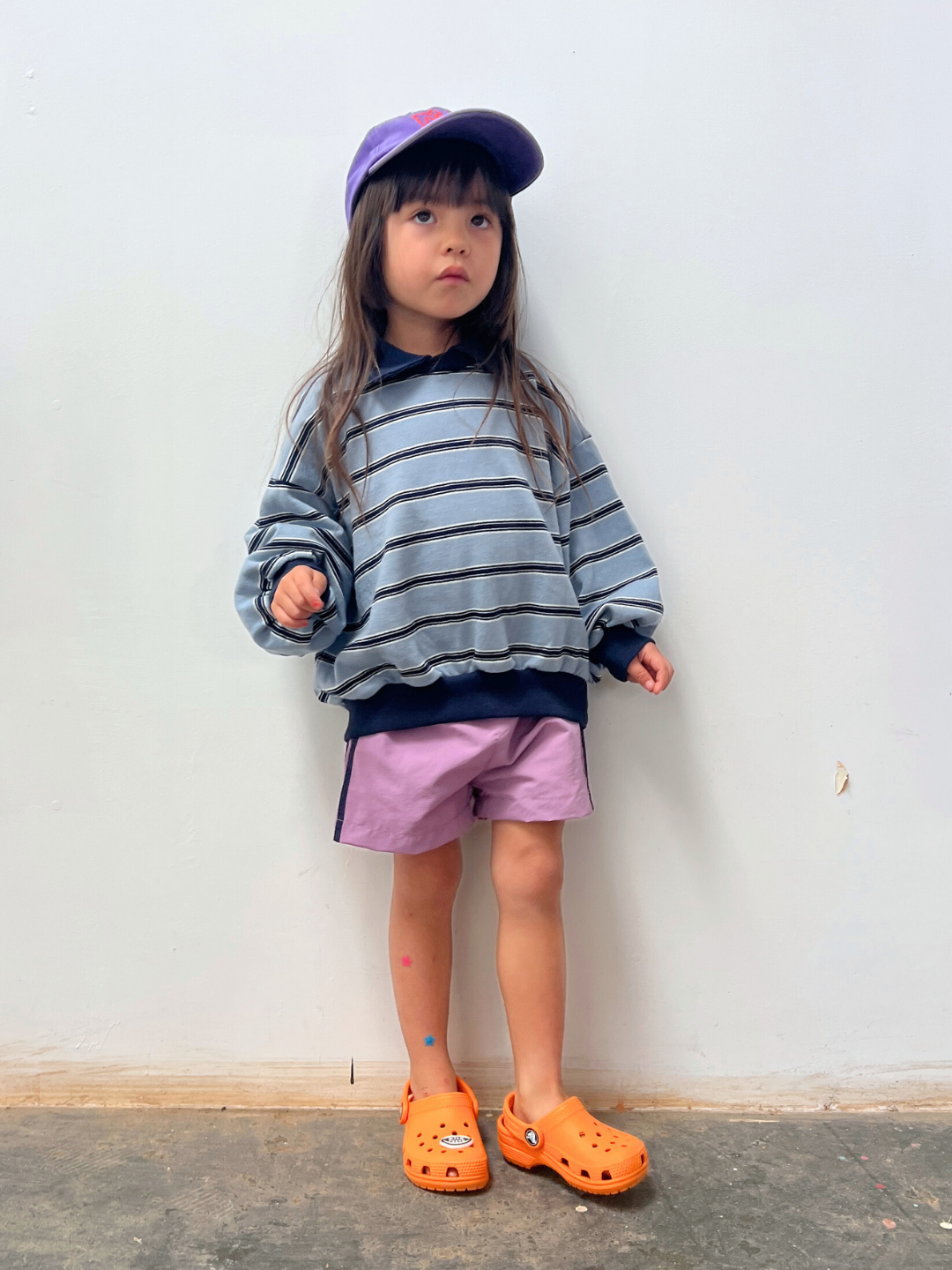 A young child stands against a plain light-colored wall wearing a GARY COLLAR SWEATSHIRT, lavender shorts, a purple cap, and cute orange animal-face clogs.