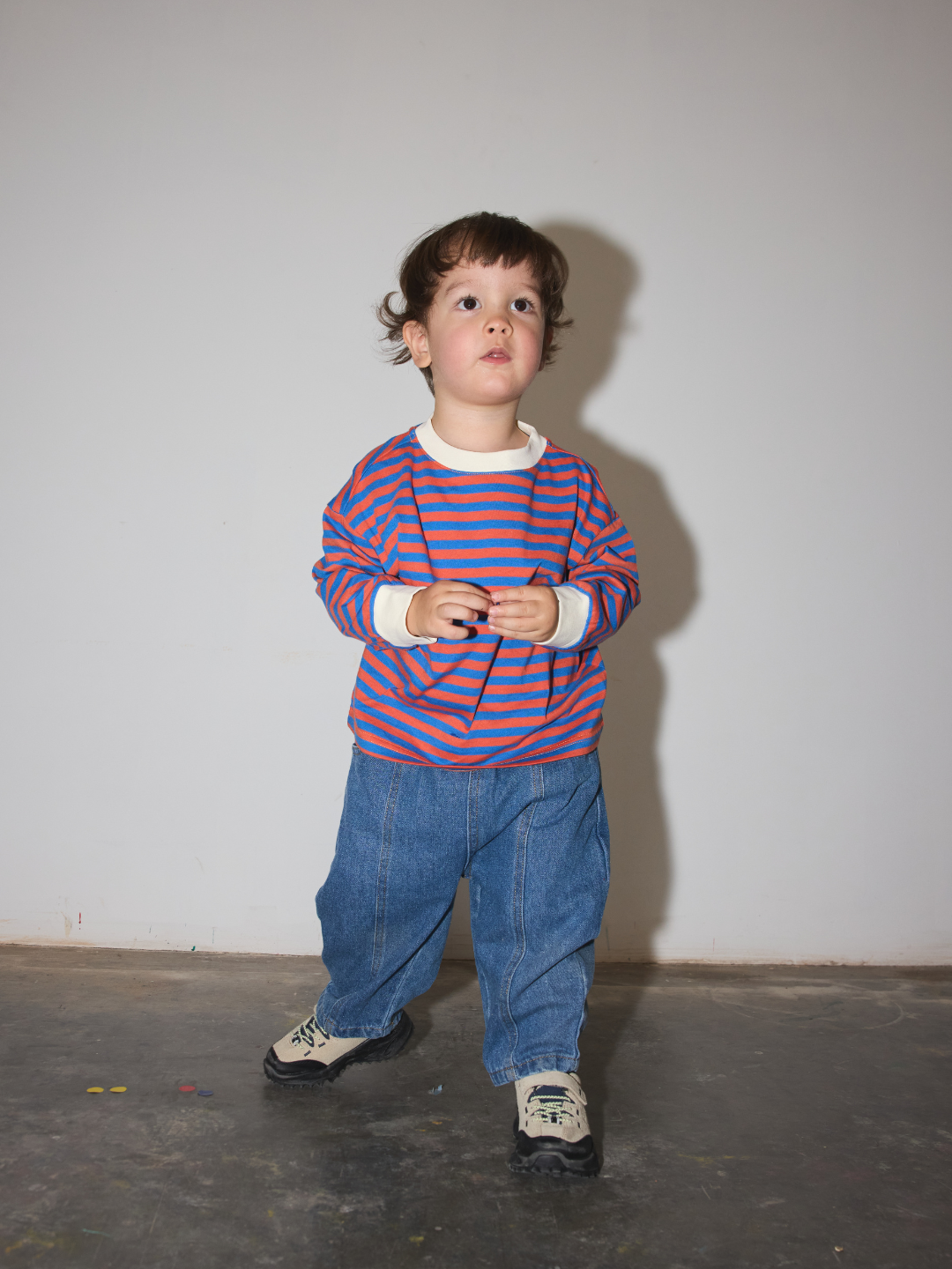 Blue | A toddler standing against a plain light gray wall, wearing a long-sleeve red and blue striped shirt with white cuffs, blue jeans, and sneakers.