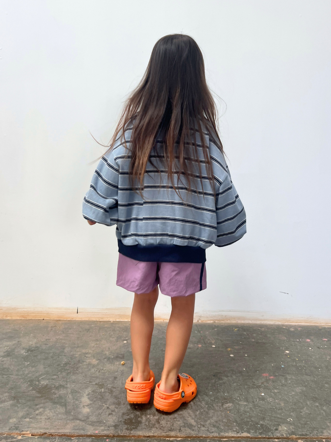 A young girl with long brown hair, wearing the GARY COLLAR SWEATSHIRT, purple shorts, and orange clogs, stands quietly facing a pale wall on a concrete floor.