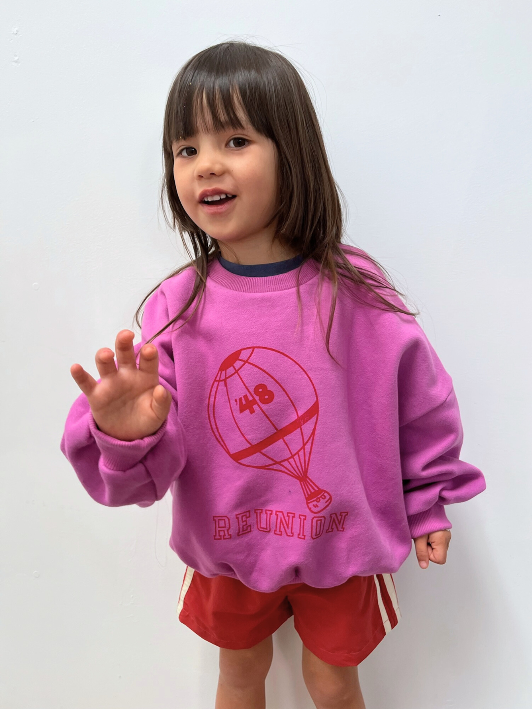 Child wearing a pink sweatshirt with a red graphic and text, standing against a white background