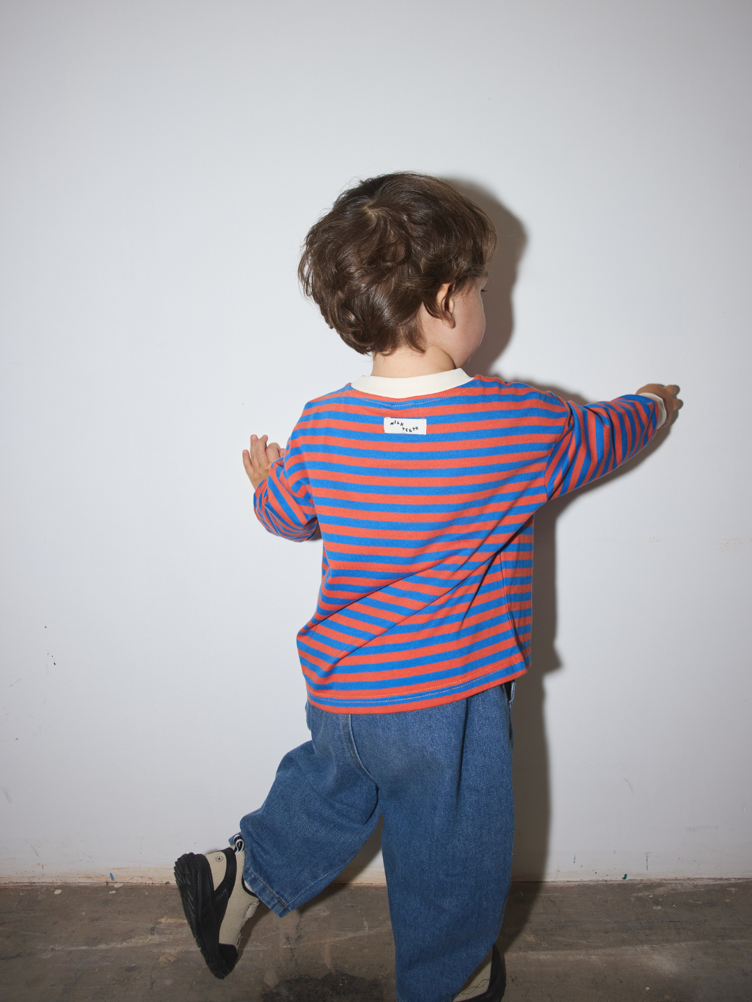 Blue | A child seen from behind wearing a red and blue horizontally striped long-sleeve shirt, blue jeans, and black sneakers, standing against a plain light gray wall.