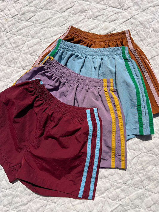 Second image of Maroon | SPRINTER SHORTS in maroon, crafted from performance fabric with an elasticated waist and blue vertical side stripes, shown on a plain white background.