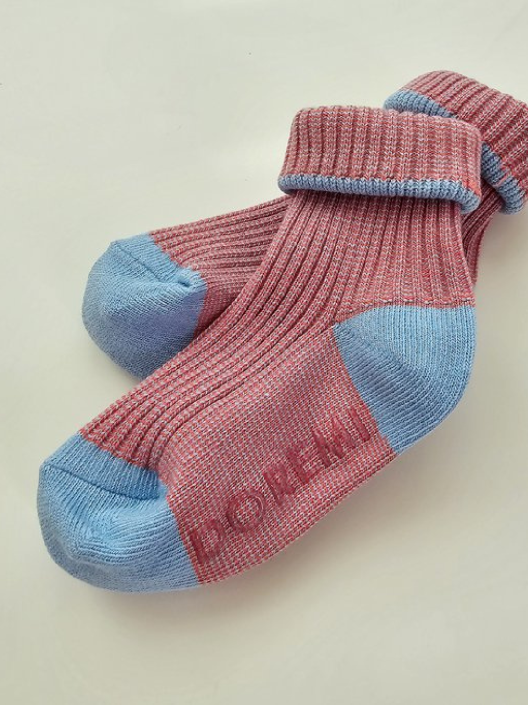 A pair of vibrant ribbed socks from the TRIO SOCK SET in pink and blue, featuring the word "DOREMI" on the sole, rests on a neutral background.