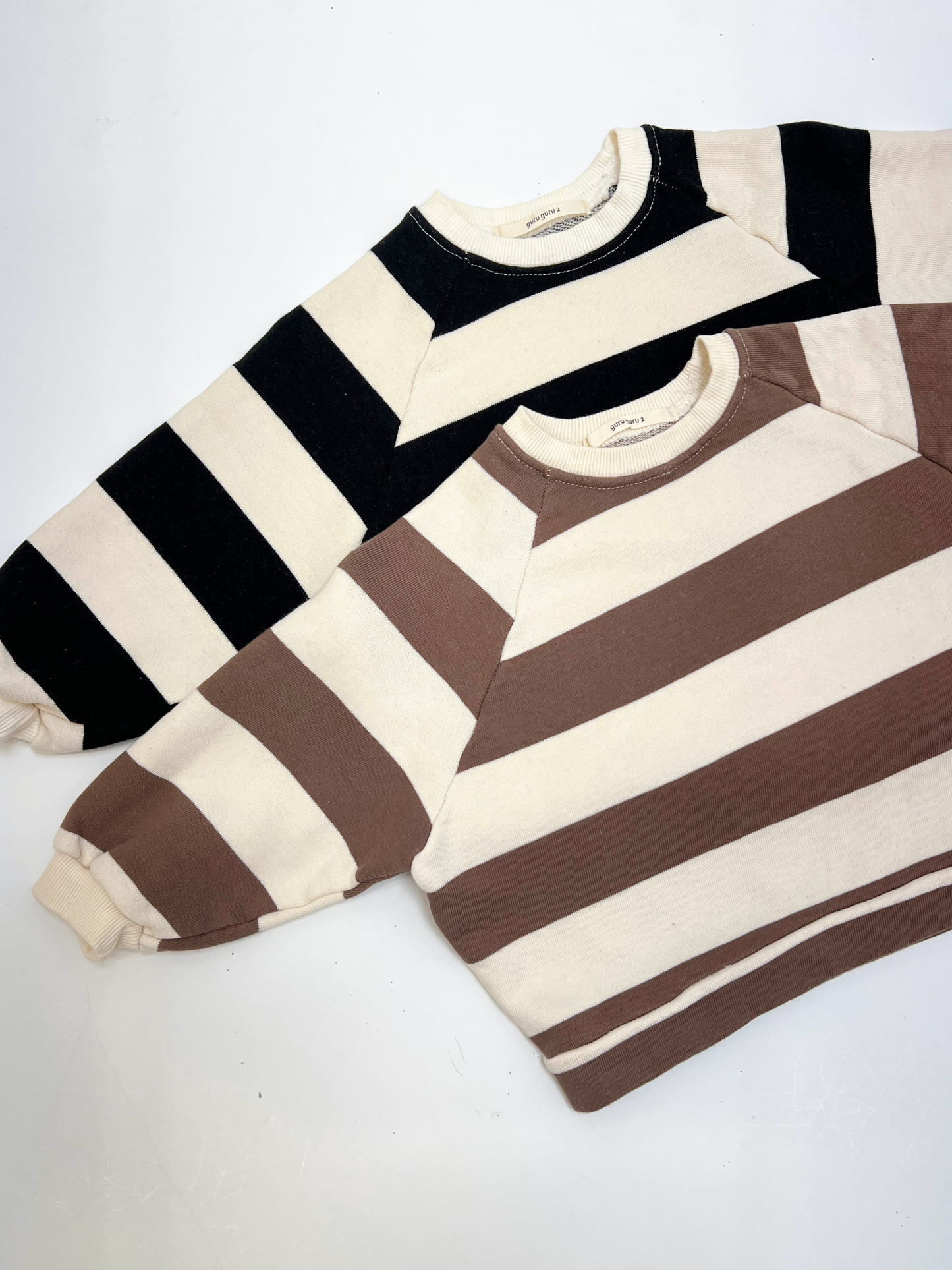 Black | Two STRIPE BABY SWEATSHIRTs from a Korean brand, made of soft cotton terry with wide black/cream and brown/cream stripes, are laid flat on a white surface.