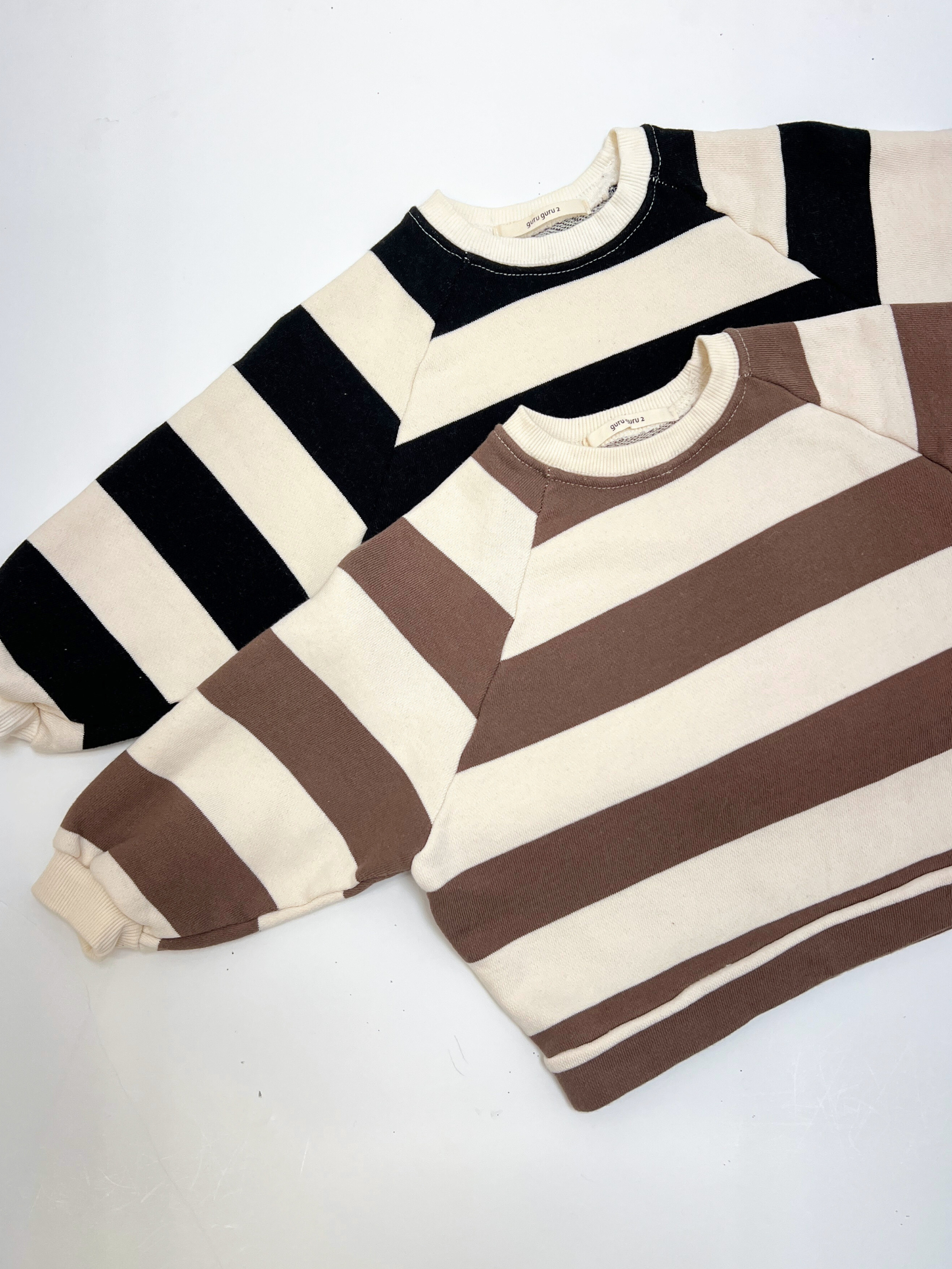 Two STRIPE BABY SWEATSHIRTs from a Korean brand, made of soft cotton terry with wide black/cream and brown/cream stripes, are laid flat on a white surface.