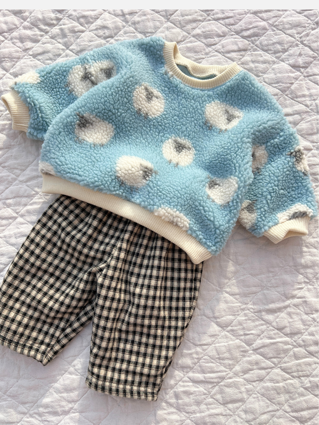 A charming SHEEP BABY SWEATSHIRT in blue with white circles, paired with black and white checkered pants, is artfully laid out on a quilted surface, capturing the essence of a beloved Korean baby brand.