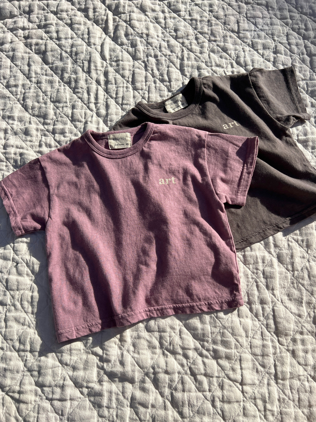 Mauve | Two kids' Studio tees laid flat on a quilt in the sun