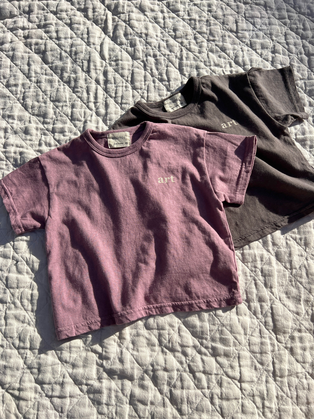 Two kids' Studio tees laid flat on a quilt in the sun