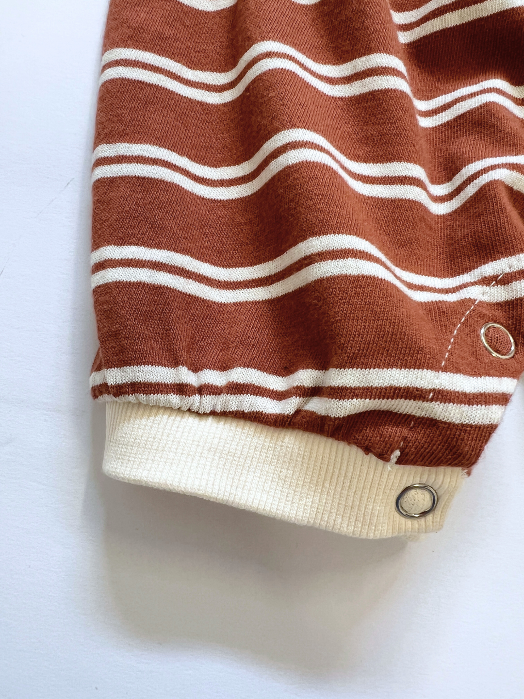 Close-up of the SLOUCHY STRIPE ROMPER’s rust cotton jersey with white stripes, a ribbed cuff, and a snap button—showcasing this Korean baby brand's romper details against a clean white background.