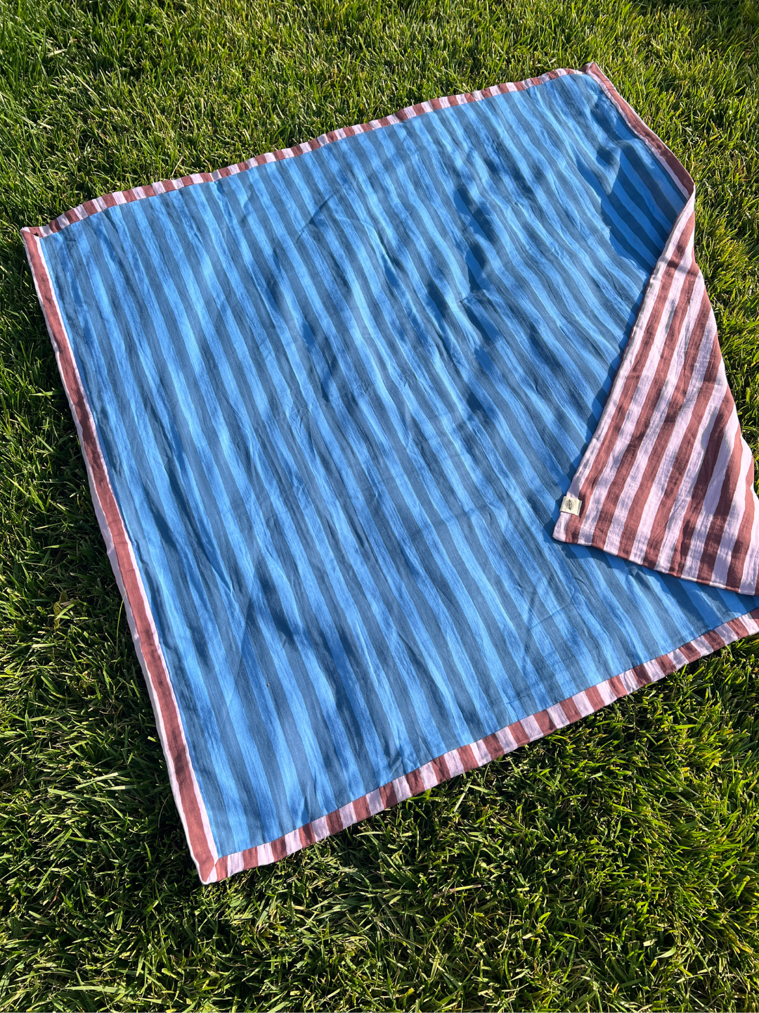 The underside of the organic cotton quilt in purple and maroon stripes, is shown on grass, with its blue striped underside showing.