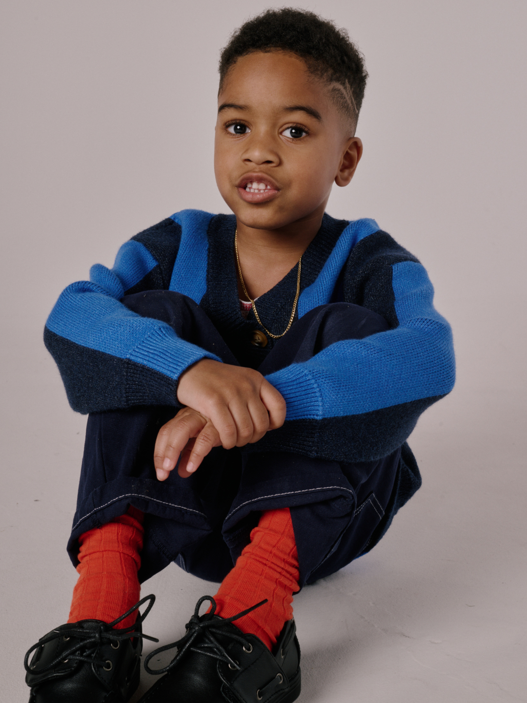 A young boy with short hair sits on the floor with his arms around his knees, wearing a STRIPE CARDIGAN, navy pants, red socks, and black shoes.