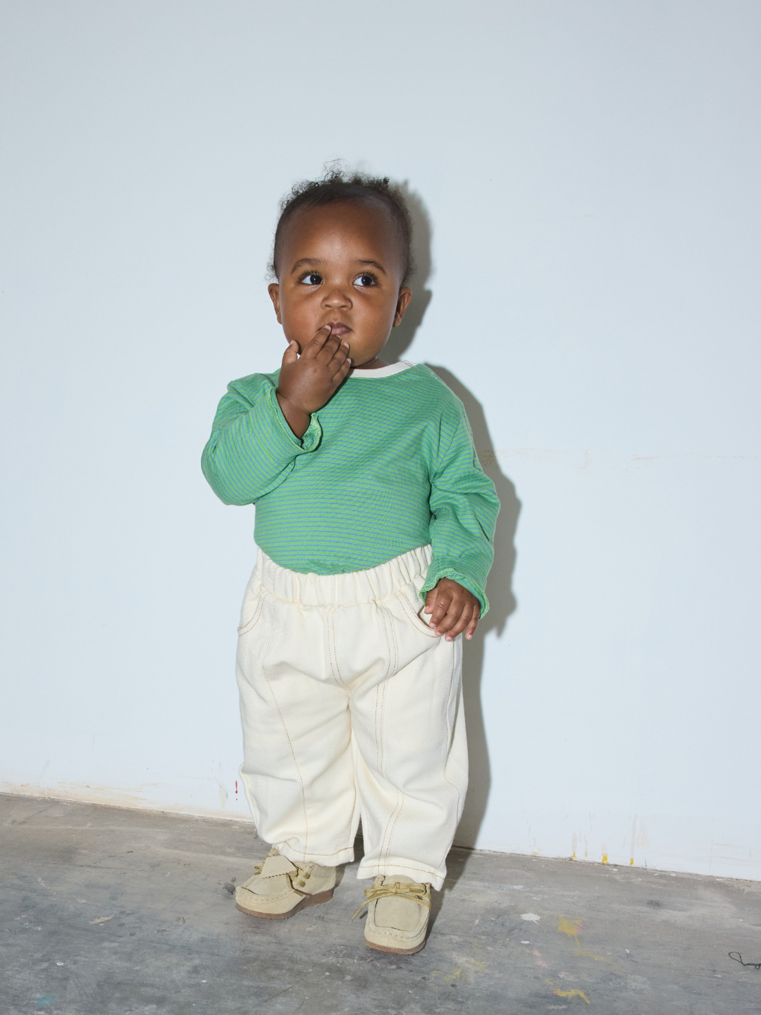 Ecru | A toddler standing against a plain light wall, wearing a green long-sleeve top, cream pants, and tan shoes.