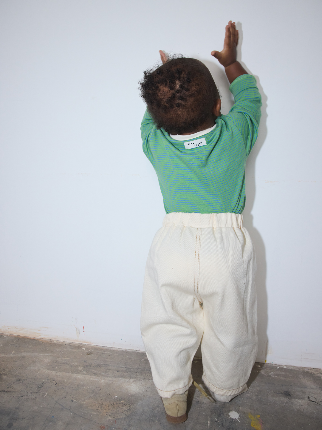 Ecru | A toddler seen from behind wearing a green long-sleeve top and cream-colored pants, standing against a plain white wall with their hands touching it.