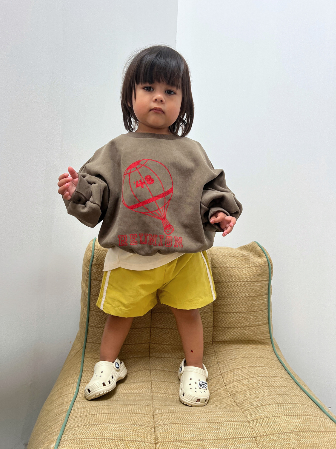Olive | Child wearing a brown sweatshirt with a red graphic and yellow shorts, standing on a beige chair against a white wall.