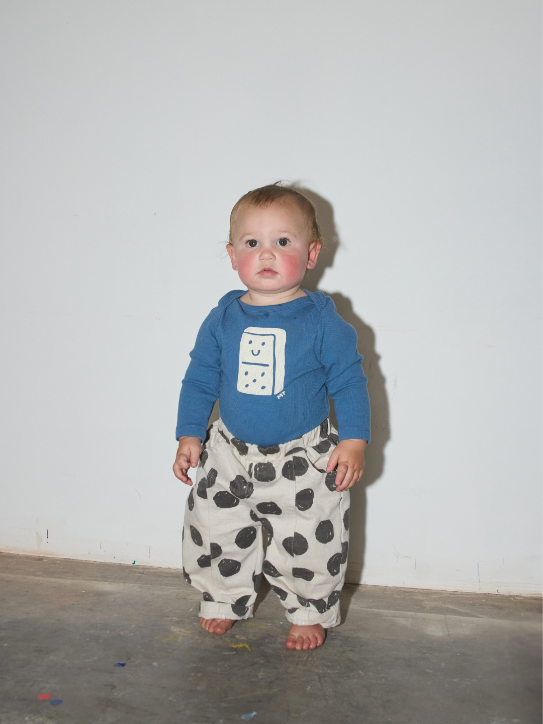 A baby standing barefoot against a plain white wall, wearing a blue long-sleeve shirt with a white domino graphic and loose white pants with black spots.