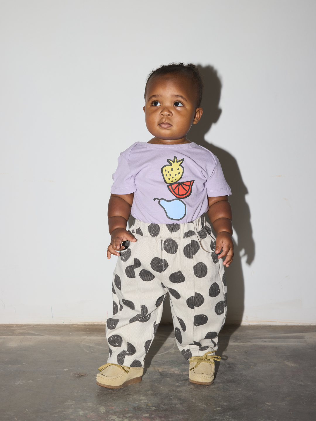 A baby standing against a plain light gray wall wearing a light purple t-shirt with a fruit graphic and white pants with black spots, paired with beige shoes.
